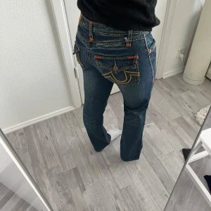 True Religion jeans - Säljer ett par True Religion Billy Super T jeans i mörkblå denim med kontrastsömmar i orange och gult. Snygga detaljer och bootcut-silhuett. Lågmidjade. 