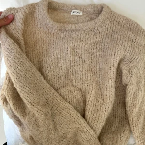 Beige stickad tröja från American Vintage - Supermjuk och chunky stickad tröja i beige från American Vintage. Tröjan har en rund halsringning, lång ärm och en mysig, lite fluffig struktur. Perfekt för lager på lager och chill höst- och vinterstil.