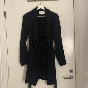 Svart kappa från Vila med bälte XS - Svart tunn kappa från Vila i storlek XS med snygg vattenfalls-krage och knytbälte i midjan. Långärmad och i en klassisk, stilren modell som passar perfekt till lager-på-lager. Enkel att matcha och ger en clean look. (Aldrig använd)