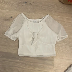 Vit blus med puffärm från Lindex - Supersöt vit blus från Lindex med korta puffärmar i transparent tyg och knytning framtill. Blusen har en croppad passform och resår i halsringningen för en bekväm känsla. Perfekt för dig som gillar romantiska och luftiga plagg.