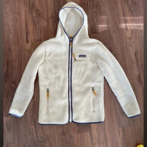 Vit teddyjacka från Patagonia - Säljer en vit teddyjacka från Patagonia med huva och blå detaljer. Jackan har dragkedja framtill, tre fickor med dragkedja och mysigt fleece-material. Perfekt för kyliga dagar och har en clean, sportig look.