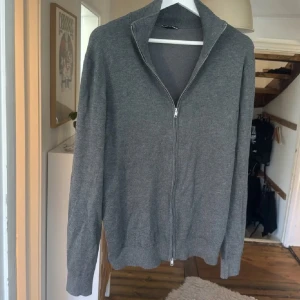 Grå kofta med dragkedja - Säljer en stilren grå kofta med hel dragkedja framtill och hög krage. Tröjan är långärmad och har en klassisk, enkel design som passar perfekt till lager-på-lager. Materialet känns mjukt och är troligtvis bomull eller en bomullsblandning.