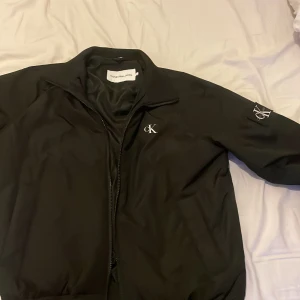 Svart Calvin Klein bomberjacka - Svart bomberjacka från Calvin Klein Jeans med klassisk cK-logga på bröstet och ärmen. Jackan har dragkedja framtill, ståkrage och slät insida. Snygg och stilren modell som passar perfekt till streetwear eller en clean look.