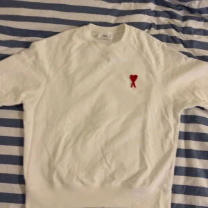 Vit sweatshirt från AMI med rött hjärta - Säljer en stilren vit sweatshirt från AMI med deras ikoniska röda hjärta och A broderat på bröstet. Tröjan har rund halsringning, långa ärmar och är gjord i mjuk bomull. Perfekt för en clean och trendig look. Står L men sitter mer som M