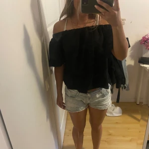 Svart offshoulder blus med puffärm - Svart blus med bred resår upptill som gör att den kan bäras offshoulder. Toppen har korta puffärmar och en luftig, lite croppad passform. Perfekt till jeansshorts eller kjol för en chill sommarlook.