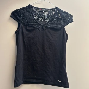 Svart spetsdetaljerad topp från Guess - Svart ärmlös topp från Guess med snygga spetsdetaljer över axlar och bröst. Toppen har v-ringning och en liten metallogo nertill. Perfekt för dig som gillar en stilren look med lite extra edge.