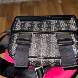 Svart axelväska från Lacoste med mönster - Snygg svart axelväska från Lacoste med geometriskt vitt mönster och tydlig logga framtill. Väskan har justerbar rem, två stora metallspännen och dragkedja under locket. Tillverkad i slitstarkt syntetmaterial med detaljer i skinnimitation.