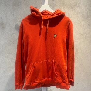 Orange hoodie från Lyle & Scott L - Snygg orange hoodie från Lyle & Scott med klassisk gul fågel-logga på bröstet. Tröjan har huva med snörning, stor magficka och ribbade muddar. Perfekt för dig som gillar streetwear och vill sticka ut med färg. Passar chill dagar och häng med kompisar.