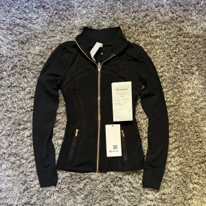 Lululemon Define Jacket - Säljer en svart slim träningsjacka från Lululemon med snygga guldiga dragkedjor och figurnära passform. Jackan har hög krage, långa ärmar och två fickor med dragkedja. Perfekt för dig som vill ha en sportig och stilren look.