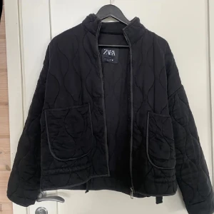 Svart quiltad jacka från Zara - Quiltad svart jacka från Zara med oversized passform och stora fickor framtill. Jackan har dragkedja och hög krage, perfekt för lager-på-lager. Snygg vågig quiltning och lite boxig siluett som ger en cool vibe.