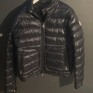Svart Moncler dunjacka med dragkedja - Snygg svart dunjacka från Moncler med klassisk quiltad design och hög krage. Jackan har två dragkedjeförsedda fickor, Moncler-logga på ärmen och stilren passform. Perfekt för lite varmare dagar där du inte anser att du behöver en tjock jacka då den är väldigt tunn