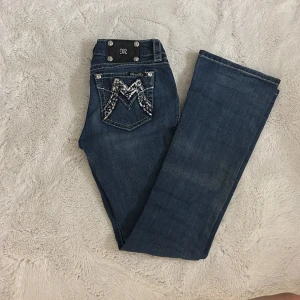 Miss Me Signature Bootcut Jeans 26 - Snygga blå bootcut jeans från Miss Me med coola detaljer och bling på bakfickorna. Klassisk femficksmodell med kontrastsömmar och dekorativa knappar. Jeansen har en normal passform och är tillverkade i slitstarkt denimtyg.