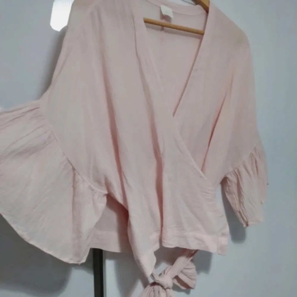 Säljer en söt ljusrosa omlottblus med knytning framtill. Blusen har trekvartsärmar med volangdetaljer som ger en romantisk touch. Perfekt för vår och sommar!. Yläosat.