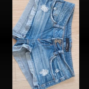 Blå jeansshorts med slitningar - Snygga blå jeansshorts med slitningar och fransiga kanter. De har en låg midja och två knappar framtill. Perfekta för en avslappnad stil.