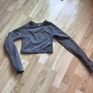 Grå croppad tränings tröja - Säljer en grå croppad tröja med lång ärm och tumhål. Tröjan är tillverkad i ett stretchigt material med 90% polyamid och 10% elastan. Perfekt för träning!