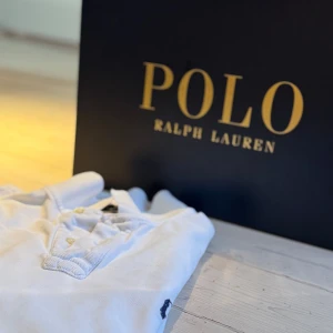 Vit pikétröja från Ralph Lauren - Säljer en klassisk vit pikétröja från Ralph Lauren med korta ärmar och den ikoniska loggan broderad på bröstet. Perfekt för en stilren och avslappnad look. Tröjan har en knappslå med tre knappar och en mjuk krage.