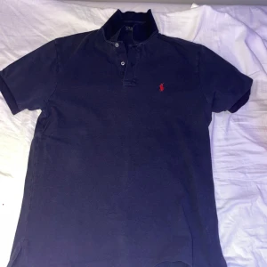 Svart kortärmad pikétröja från Ralph Lauren - Snygg svart pikétröja från Ralph Lauren med klassisk krage och knappar. Den har en röd broderad logotyp på bröstet. Perfekt för en stilren look. Den är i storlek s passar perfekt och bra skick 