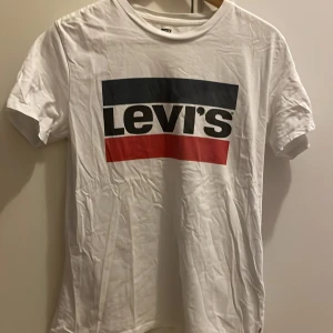 Vit t-shirt från Levi's - Säljer en klassisk vit t-shirt från Levi's med deras ikoniska logga i svart, blått och rött på bröstet. T-shirten är kortärmad och gjord i mjuk bomull. Perfekt för en avslappnad stil.