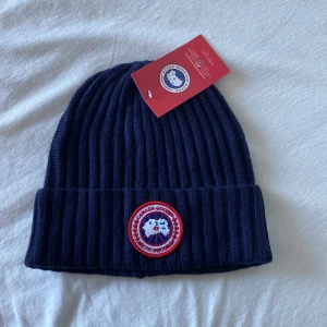 Mörkblå ribbad mössa från Canada Goose - Snygg mörkblå ribbad mössa från Canada Goose med deras ikoniska märke framtill. Perfekt för kyliga dagar och håller dig varm med stil. Mössan har en klassisk design och är ett måste för vintern.