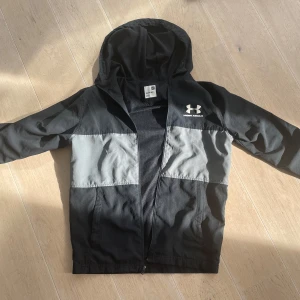 Svart och grå vindjacka från Under Armour - Säljer en snygg svart och grå vindjacka från Under Armour. Jackan har en dragkedja framtill och en praktisk huva. Perfekt för blåsiga dagar och träning utomhus. 🖤 dragkedjan fungerar ej så bra , pga att jag säljer biligt 