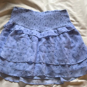 Blå blommig kjol från Gina Young  - Söt blå kjol från Gina Tricot med blommigt mönster och volangdetaljer. Den har en elastisk midja för en bekväm passform. Perfekt för en somrig look!