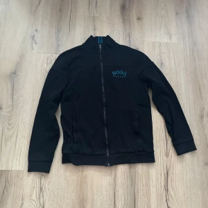 Svart tröja med dragkedja från Hugo Boss - Säljer en stilren svart tröja från Hugo Boss med dragkedja och blå logga på bröstet. Perfekt för en casual look. Tröjan har långa ärmar och en hög krage.