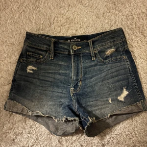 Blå jeansshorts från Hollister - Snygga blå jeansshorts från Hollister med hög midja och slitna detaljer. De har en klassisk femficksdesign och fransiga kanter för en avslappnad look. Perfekta för sommardagar!
