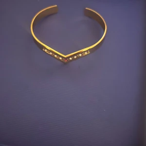 Guldarmband med stenar - Elegant guldarmband med en V-formad design. Armbandet är dekorerat med glittrande stenar, inklusive en röd sten i mitten. Perfekt för att ge en lyxig touch till din stil.❤️