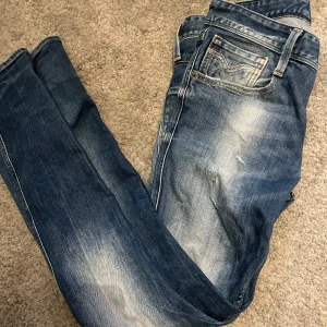 Blå jeans från Replay - Snygga blå jeans från Replay med en klassisk femficksdesign och diskreta slitningar för en trendig look. De har en knappgylf och är perfekta för en avslappnad stil. Storlek 28 32