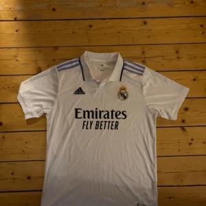 adidas Real Madrid 2022 - 23 Home Jersey - White / Light Purple - Aldrig använd så alltså helt ny, Säljer en vit Real Madrid fotbollströja från Adidas med korta ärmar. Tröjan har klubbens emblem och Adidas-logga på bröstet samt sponsortryck 'Emirates Fly Better'. Den har en klassisk krage med svarta detaljer och tre ränder i lila på axlarna.
