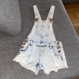Ljusa jeansshorts med hängslen från Dungaree, storlek 34 - Snygga ljusa jeansshorts med hängslen från Dungaree. De har slitna detaljer och fransar vid benen. Knappar i sidorna och en praktisk bröstficka. Perfekta för en avslappnad stil.