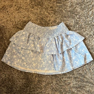 Blå blommig kjol med volang - Söt blå kjol med blommigt mönster och volangdetaljer. Den har en elastisk midja för bekväm passform. Perfekt för en somrig look! Bara använd en gång!! 💕