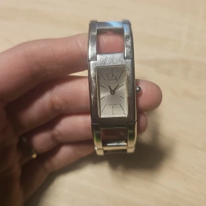 Silverfärgad klocka från Calvin Klein - Elegant silverfärgad klocka från Calvin Klein med rektangulär urtavla och stilren design. Armbandet är i metall med öppna länkar som ger en modern look. Perfekt för dig som gillar minimalistisk stil. Kostar ca 3000 som ny