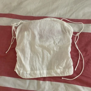 Vit axelbandslös sommartopp - Säljer en söt vit axelbandslös topp med knytband. Toppen har en smockad detalj på baksidan för bättre passform. Perfekt för varma sommardagar!