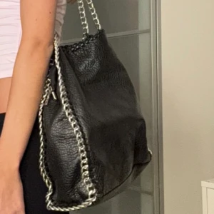 Svart axelväska med kedjedetaljer - Snygg svart axelväska i skinn med coola kedjedetaljer längs kanterna. Väskan har en praktisk dragkedja och rymlig design, perfekt för att bära med sig allt man behöver. Den stilrena looken gör den lätt att matcha med olika outfits.