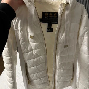 Vit quiltad jacka från Barbour - Säljer en snygg vit quiltad jacka från Barbour med dragkedja och huva. Jackan har en stilren design med Barbour-logga på ärmen och praktiska fickor med dragkedjor. Perfekt för kyligare dagar.