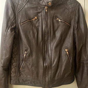  skinnjacka Rock N Blue brun med dragkedjor(rosè) - Snygg brun skinnjacka med flera dragkedjor och quiltade detaljer på axlarna. Jackan har en hög krage med knapp och dragkedjor vid ärmsluten för extra stil. Perfekt för en cool look!