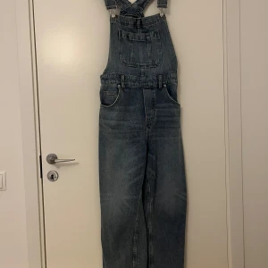 Blå hängselbyxor  - Snygg blå hängselbyxor med justerbara axelband och flera fickor fram och bak. Byxdressen har knappar på sidorna för enkel på- och avtagning. Perfekt för en avslappnad stil.