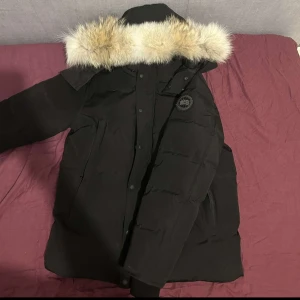 Canada Goose Helt ny - Säljer en svart parkajacka med en snygg pälsdetalj på huvan. Jackan har långa ärmar och knappar framtill. Perfekt för kalla dagar när du vill hålla dig varm och stilren. Öppen för byten!