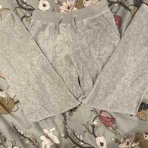 Grå mjukisbyxor från Juicy Couture - Snygga grå mjukisbyxor från Juicy Couture med broderad logga och snörning i midjan. Byxorna har praktiska fickor med knappar baktill och är perfekta för en avslappnad stil.