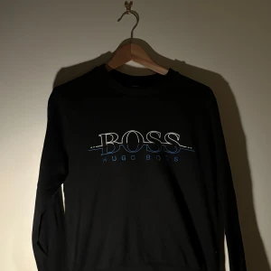 Svart tröja från Hugo Boss - Snygg svart tröja från Hugo Boss med deras ikoniska logga i vitt och blått på framsidan. Tröjan har långa ärmar och en klassisk rund halsringning. Perfekt för en stilren look.