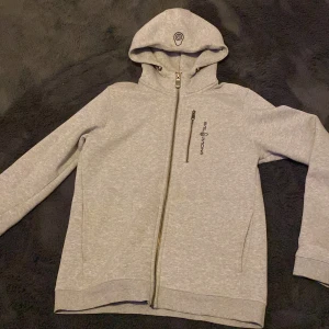 Grå huvtröja från Sail Racing - Säljer en grå huvtröja från Sail Racing med dragkedja och justerbar huva. Tröjan har långa ärmar och en stilren design med logotyp på bröstet och huvan. Perfekt för en avslappnad stil. Den är använd ett fåtals gånger men är som ny. 