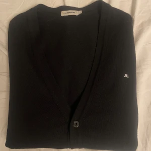 J.Lindeberg Kofta - Säljer en stilren svart J.Lindeberg kofta med knappar framtill. Koftan har en v-ringad design och långa ärmar, perfekt för en avslappnad look. Passar bra till jeans eller chinos. Den är i mycket bra skick👍