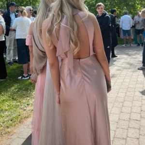 Balklänning  - Elegant rosa långklänning med öppen rygg och knytband i nacken. Klänningen har en slät design och är perfekt för festliga tillfällen. Den är ärmlös och har en vacker glansig yta som ger en lyxig känsla. Nypris 1799kr