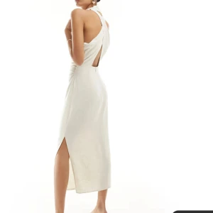 Beige klänning från ASOS - Elegant beige klänning från ASOS med hög hals och öppen rygg. Klänningen har en smickrande slits och är ärmlös, vilket ger en stilren look. Perfekt för en chic och modern stil. Aldrig använt !