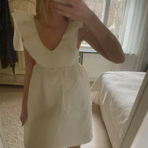 Studentklänning  - Säljer en söt vit klänning med volangdetaljer och v-ringning. Klänningen har en kort längd och är perfekt till studenten. Materialet har en fin textur som ger en extra touch.