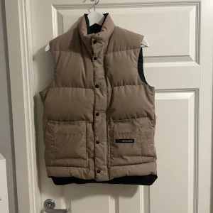 Beige dunväst från Canada Goose - Säljer en snygg beige dunväst från Canada Goose. Västen har en hög krage och knäppning med knappar framtill. Två stora fickor på framsidan ger både stil och funktion. Perfekt för kyliga dagar! XS men sitter som S/M
