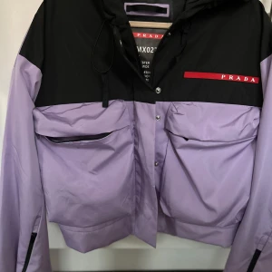 Lila och svart jacka från Prada - Snygg lila och svart jacka från Prada med dragkedja och knappar. Jackan har en modern design med två stora fickor framtill och en stilren logga på bröstet. Perfekt för att ge din outfit en lyxig touch.