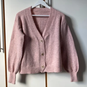 Rosa kofta med knappar - Mysig rosamelerad kofta med v-ringning och fina knappar framtill. Den har långa ärmar, lite puffade nedtill med smal mudd. Helt oanvänd. Längd 60 cm. 