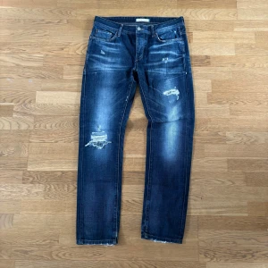 Mörkblå jeans från Jack & Jones - Snygga mörkblå jeans från Jack & Jones Vintage Co. med slitna detaljer och en klassisk femficksdesign. Perfekta för en avslappnad stil med en touch av vintage. Märkeslapp baktill och knappgylf framtill.